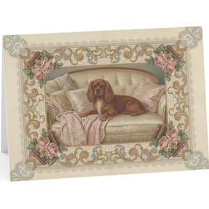 May include: A greeting card depicting a Cavalier King Charles Spaniel dog on a cream sofa with pink cushions and a blanket. The card has a decorative border with roses and scrollwork.