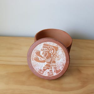 May include: A terracotta-colored ceramic container with a matching lid. The lid features a white and terracotta-colored carved design of a figure. The container is open, revealing its interior.