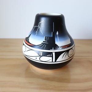 May include: A black ceramic vase with a unique design. The vase features a landscape scene with a lightning bolt, a building, and a white and orange border. The vase is on a wooden surface.