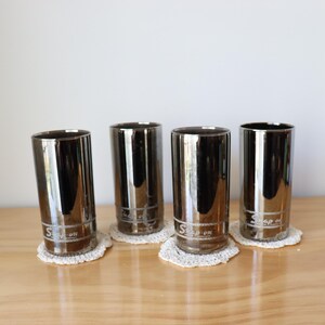 May include: Four chrome-plated drinking glasses with a reflective surface, each resting on a small, white, crocheted doily. The glasses have a cylindrical shape and a logo near the base. They are arranged on a light-colored wooden surface.