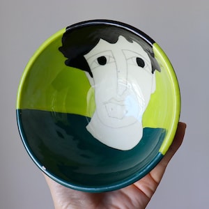 May include: A ceramic bowl with a hand-painted face design. The bowl features a lime green interior, a dark teal exterior, and a black rim. The face is depicted with a white base and black details. A unique piece for serving or display.