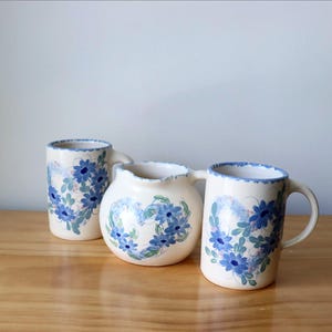 May include: Three ceramic pieces: a cylindrical mug, a round pitcher, and a mug with a handle. All are white with blue floral patterns and scalloped edges. The pitcher has two handles. The items are on a wooden surface.