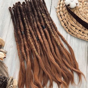May include: A bundle of brown and auburn dreadlocks, with a gradient effect. The dreadlocks are adorned with gold-colored thread and small black beads. A straw hat with a brown band and cotton accents is in the background.