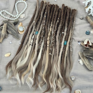 May include: A bundle of brown and blonde dreadlocks with decorative white thread and turquoise beads. The dreadlocks have a gradient effect, transitioning from dark brown to blonde. The image is taken from above, with seashells and pearls in the background.