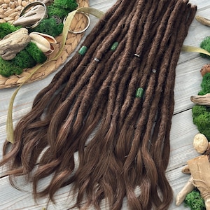 May include: Dark brown dreadlocks with lighter brown ends, adorned with green and silver accents. The dreadlocks are arranged on a decorative woven mat with moss and natural elements, creating a bohemian aesthetic.