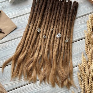 May include: A set of brown and blonde dreadlocks with silver charms. The dreadlocks are arranged on a light blue wooden surface, with wheat stalks and a small white box in the background. The dreadlocks have a gradient color effect.