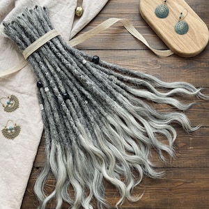 Puede incluir: Un conjunto de rastas sintéticas ombre grises y blancas con cuentas negras. Las rastas están dispuestas en un estilo suelto y ondulado y están atadas juntas con una cinta marrón.