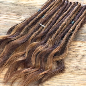 May include: A collection of brown and auburn dreadlocks with wavy ends. Some dreadlocks have small silver charms and blue beads attached. The dreadlocks are arranged on a wooden surface, showcasing the varying shades of brown and auburn.