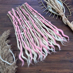 May include: A set of synthetic dreadlocks featuring a blend of light pink, beige, and off-white strands. The dreadlocks are textured with various wraps, small white beads, and metallic accents, transitioning into loose, wavy ends at the bottom.