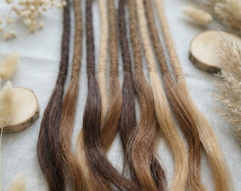 Op de afbeelding: Een set van tien haarextensies met gevlochten bovenkanten die overgaan in losse, golvende uiteinden. De extensies tonen een verloop van natuurlijke haarkleuren, variërend van donkerbruin tot lichtblond, gerangschikt in een rij.