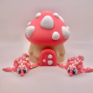 May include: A whimsical mushroom house with a red cap and white spots, a tan base, and a pink door. Two pink and white creatures with blue eyes are positioned on either side of the house, creating a fantasy scene.