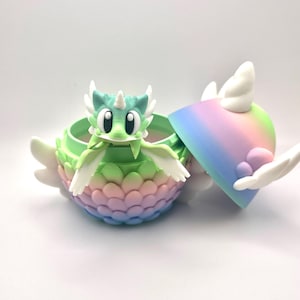 May include: A whimsical, colorful dragon figurine inside a matching container. The dragon is green with white accents and a small horn. The container is a gradient of pastel colors, with a lid that is partially open, revealing the dragon.