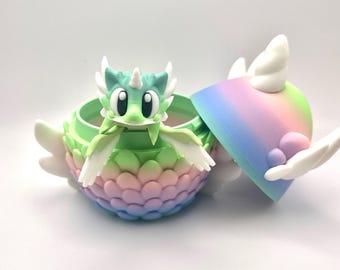 May include: A whimsical, colorful dragon figurine inside a matching container. The dragon is green with white accents and a small horn. The container is a gradient of pastel colors, with a lid that is partially open, revealing the dragon.