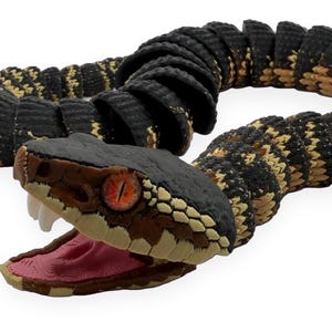 May include: A segmented articulated snake toy featuring a black and tan patterned body. The head is black with brown accents, featuring bright orange eyes with vertical pupils and an open mouth revealing white fangs and a pink interior.