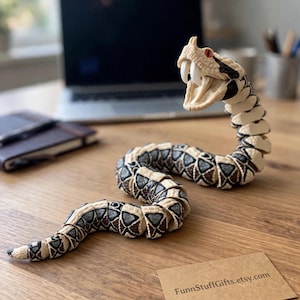May include: An articulated snake figurine with a cream-colored body, dark brown and black diamond-shaped patterns, and red eyes. The snake has an open mouth with two prominent white fangs. A small brown card displays the text FunnStuffGifts.etsy.com.