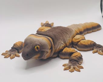 May include: A detailed, realistic sculpture of a gecko in shades of brown, tan, and orange. The reptile is lying flat, with its legs spread out. The gecko has textured skin, a detailed head, and yellow eyes. The overall design is lifelike.