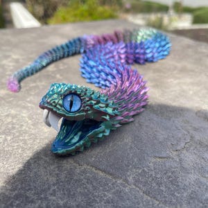 May include: A iridescent purple, blue, and green snake figurine with a blue eye and an open mouth. The snake is curled up and has a scaly texture.