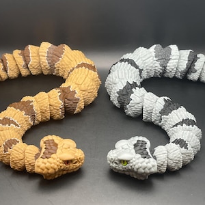 May include: Two articulated snake toys. One is tan and brown, the other is gray and black. Both have detailed heads with realistic features and are posed in a curved position. The snakes are made of a durable material.