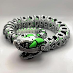 May include: A 3D-printed articulated snake toy in a coiled position. The toy features a robotic design with a color scheme of gray, white, and green. The snake's head is detailed with eye sockets and a segmented jaw. The body is made of interlocking segments.