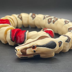 May include: A segmented, cream-colored snake toy with brown and black markings. The snake's head features a detailed design with an orange eye and a brown and black pattern. The segments near the head are red.