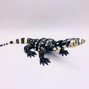 May include: A black and white lizard figurine with a spotted pattern. The reptile has a long tail and is posed on a white surface. The figurine is a detailed representation of a lizard.