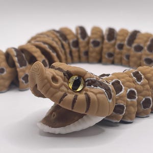 May include: A detailed, segmented snake toy in shades of brown and tan with dark brown spots. The snake's head is open, revealing a white interior. The eye is a reflective yellow color. The toy is on a white background.