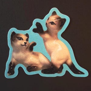 May include: A glittery sticker featuring two Siamese cats. One cat is lying down, while the other is standing and reaching out. The cats have tan fur with dark brown markings and are outlined with a light blue border.