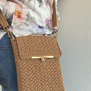 Op de afbeelding: Een bruine gehaakte schoudertas met een gouden sluiting. De tas wordt over de schouder gedragen door iemand die een bloezen met bloemenprint en een blauwe jeans draagt.