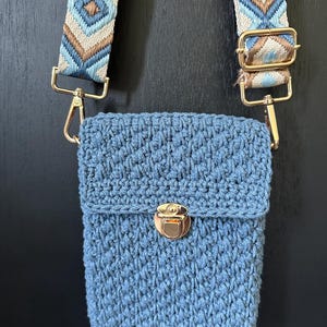 May include: A blue crochet crossbody bag with a patterned strap. The bag has a rectangular shape with a flap closure and a gold-coloured clasp. The strap features a geometric design in shades of blue, tan, and white.