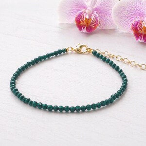 May include: A delicate bracelet featuring small, faceted dark green beads strung together. The bracelet includes a gold-toned metal lobster claw clasp and a short adjustable chain extension, providing a secure and flexible fit for the wearer.