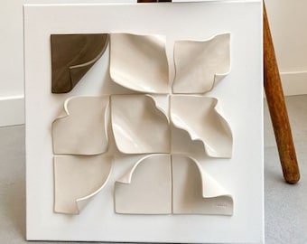 May include: A white canvas with nine ceramic tiles arranged in a grid pattern. The tiles are all white except for one, which is brown. The tiles are all slightly raised and have a textured surface. The canvas is hanging on a wall.