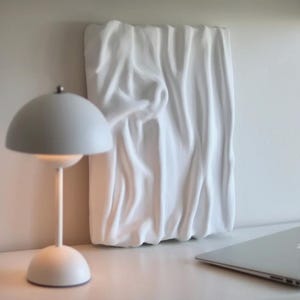 Puede incluir: Un colgante de pared blanco texturizado con un diseño de tela drapeada. Una lámpara de escritorio blanca con una pantalla redondeada y un ordenador portátil están sobre una superficie blanca. La lámpara tiene una base pequeña y redonda y un tallo delgado.