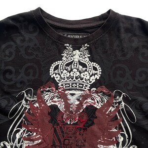 May include: Black t-shirt with a large white and red graphic design featuring a crown, wings, and ornate details. The shirt has a crew neck and a subtle pattern in the background. The brand name "AVIREX" is visible on the tag.