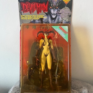 May include: A red and black action figure of Devilman Lady from the Devilman anime series. The figure is posed with her arms outstretched and her legs slightly bent. The figure is packaged in a clear plastic blister pack with a red and black cardboard backing. The packaging features the Devilman logo and the text "Special Bonus Part Included! You Can Collect All of 8 Figures! Display Base Included!" The packaging also features a glow-in-the-dark feature.