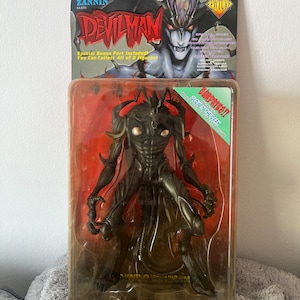 May include: A Devilman action figure in its original packaging. The figure is dark gray with a detailed, demonic design. The packaging includes the text "DEVILMAN" and "ZANNIN", along with promotional text. The figure is in a clear plastic case.
