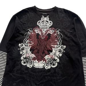 May include: Black long-sleeve shirt with a large, ornate crest design in burgundy and white. The crest features a crown, eagle, and scrollwork. The sleeves have a striped pattern. The shirt has the word "AVIREX" printed on the collar.