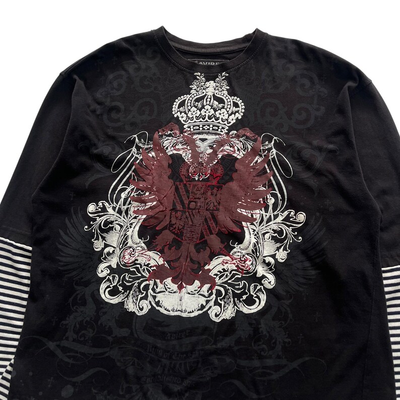 May include: Black long-sleeve shirt with a large, ornate crest design in burgundy and white. The crest features a crown, eagle, and scrollwork. The sleeves have a striped pattern. The shirt has the word "AVIREX" printed on the collar.