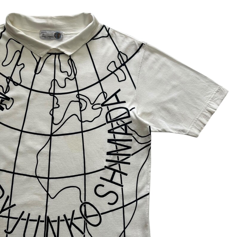 May include: Cream-colored polo shirt with a black world map design. The shirt features short sleeves and a collared neckline. The text "OSHIMADA" is printed on the front. A small tag with text is visible near the collar.