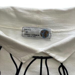 May include: Close-up of a white collared shirt with a black graphic design. The shirt features a tag with the text "MAXFLI BY JUNKO SHIMADA MADE IN JAPAN" and a globe graphic.