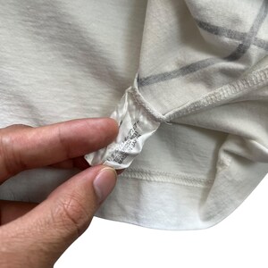 May include: Close-up of a cream-colored t-shirt with a tag attached. The tag has Japanese text and the words "MADE IN JAPAN". The shirt has a subtle grid pattern in a darker color.