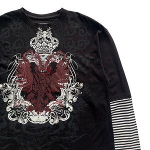 May include: Black long-sleeve shirt with a large crest design in white and burgundy. The crest features a crown, wings, and ornate scrollwork. The sleeves have a striped pattern.