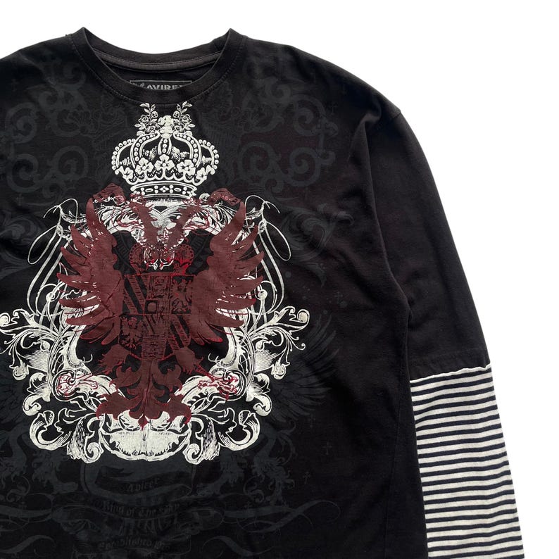 May include: Black long-sleeve shirt with a large crest design in white and burgundy. The crest features a crown, wings, and ornate scrollwork. The sleeves have a striped pattern.