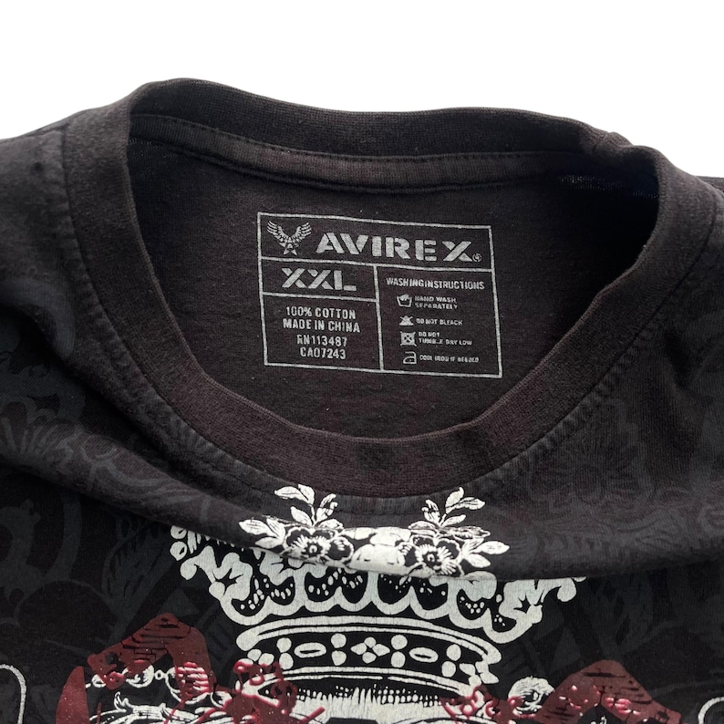 May include: Black XXL cotton t-shirt with a decorative crown design and the brand name AVIREX. The shirt has a crew neck and a tag with washing instructions and fabric details.