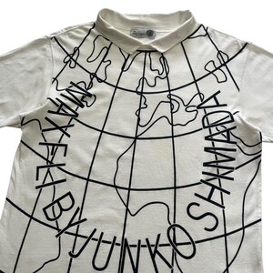May include: White collared t-shirt with a black world map design and the text "MAX BY YUNKO SHYAMADA" in a circular pattern. The shirt has short sleeves and a small tag near the collar.