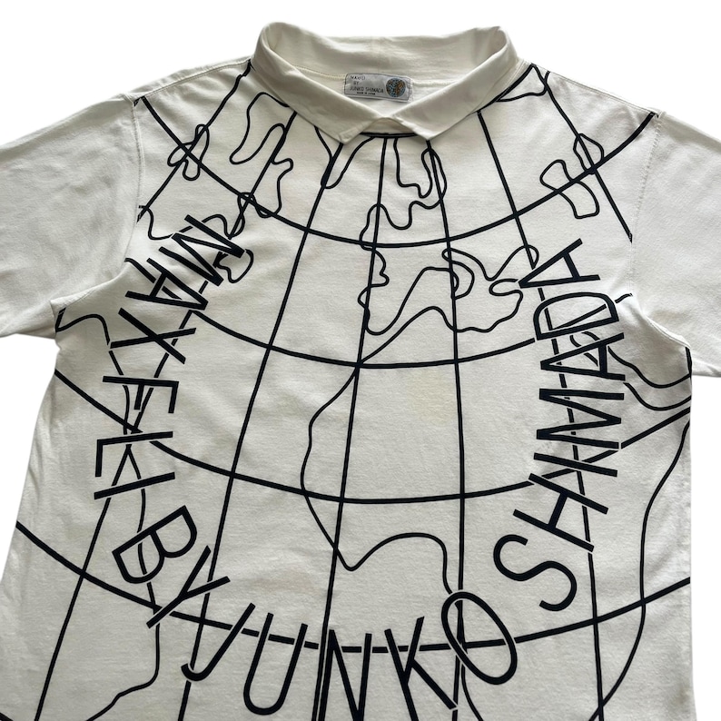 May include: White collared t-shirt with a black world map design and the text "MAX BY YUNKO SHYAMADA" in a circular pattern. The shirt has short sleeves and a small tag near the collar.