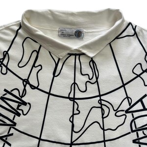 May include: Cream-colored polo shirt with a black world map design. The shirt features a white collar and the text "MAXFU BY JUNKO SHIMADA" on a label near the collar. The graphic design covers the front of the shirt.