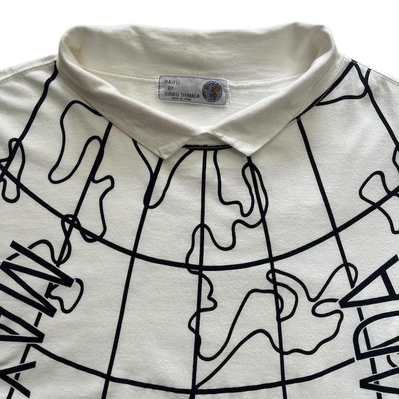 May include: Cream-colored polo shirt with a black world map design. The shirt features a white collar and the text "MAXFU BY JUNKO SHIMADA" on a label near the collar. The graphic design covers the front of the shirt.
