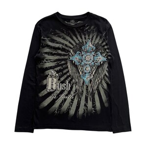 Op de afbeelding: Zwart shirt met lange mouwen met een grafisch ontwerp met een kruis, vleugels en de tekst "Rush in Faith". Het kruis heeft blauwe accenten. Het ontwerp staat tegen een grijze zonnestralen achtergrond.