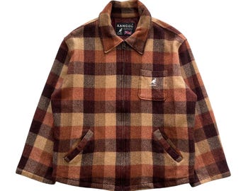 Könnte beinhalten: Eine braune, orange und beige karierte Jacke mit durchgehendem Reißverschluss. Die Jacke hat einen Kragen, zwei Vordertaschen und eine kleine Brusttasche. Der Markenname "KANGOL" ist auf dem Kragen zu sehen.