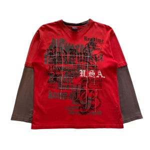 May include: Red long-sleeve shirt with a gray undershirt. The shirt features a graphic design with the word "USA" and other text. The shirt has a crew neck and is made of a soft material.